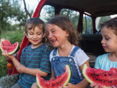 Children laughing and eating watermelon, sitting on the back of a truck.